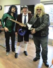 Three men with heavy metal outfits and wigs. One holding an electric guitar.